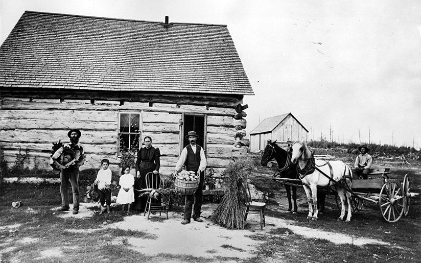 Photo of Norwegian settlers with their horses in front of a log house in the late 1800s